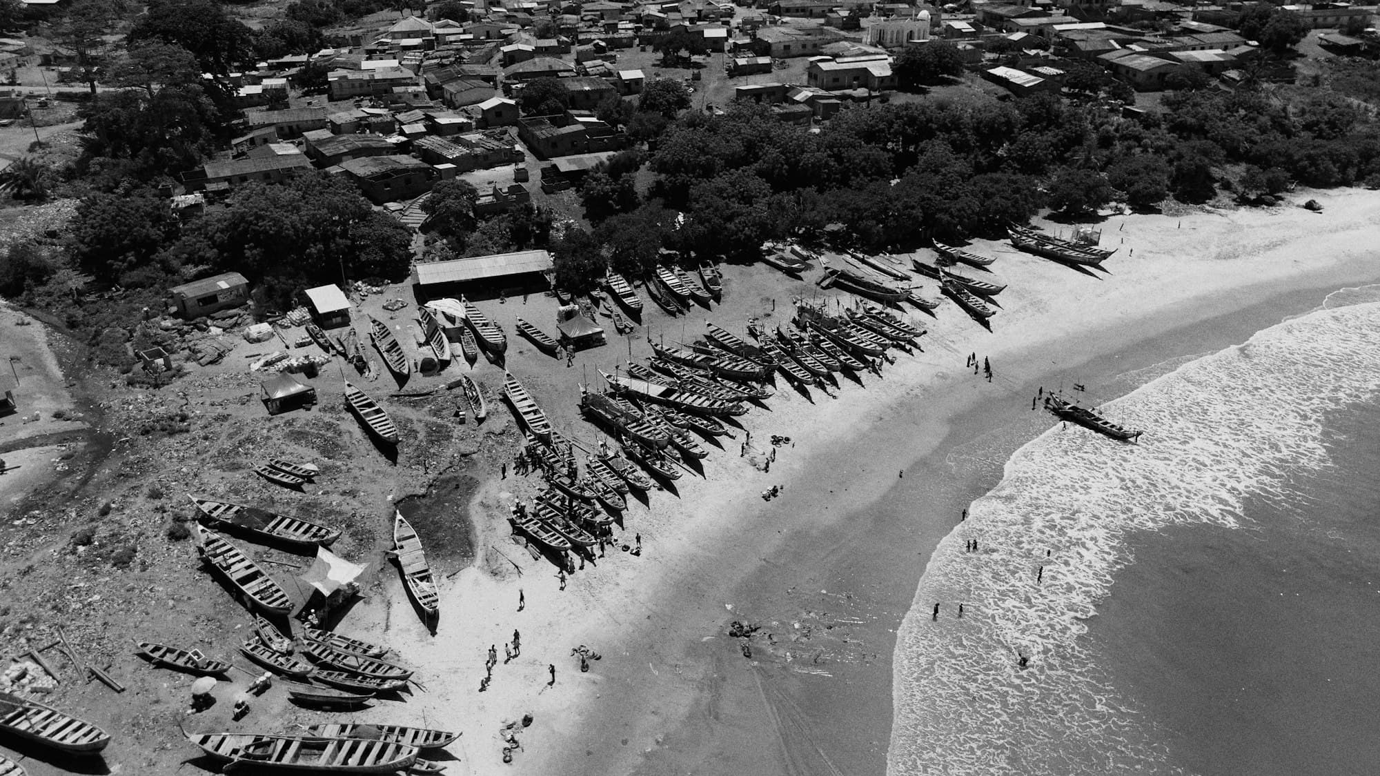 Fishing canoes drawn up on a curving beach below a coastal town, in black and white, seen from above